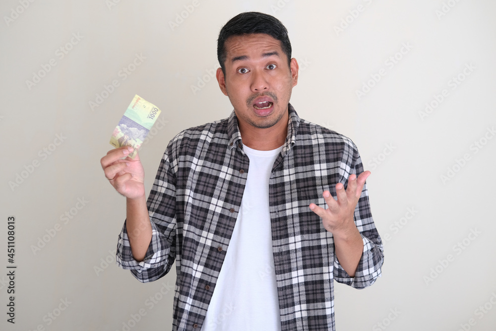 Adult Asian man holding small amount of paper money with worried face expression Adult Asian man holding small amount of paper money with worried face expression