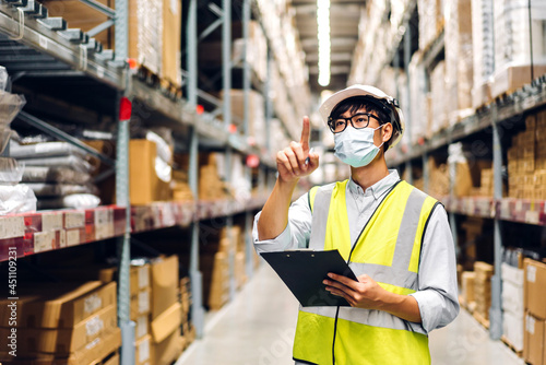 Portrait of asian engineer man in helmet in quarantine for coronavirus wearing protective mask order details checking goods and supplies on shelves with goods background in warehouse.logistic © Art_Photo