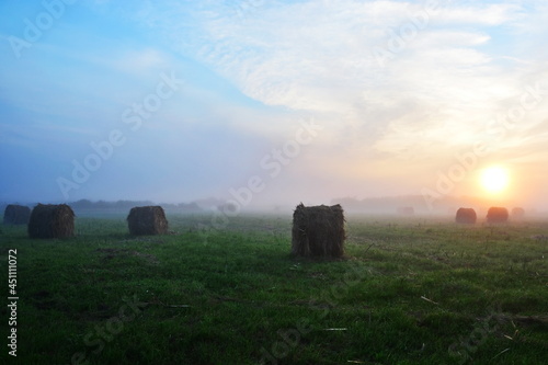 Morning rustic rural country landscape. Big field with mown grass and haystacks in the morning fog on background of blurred rising sun with blue gradient in sky
