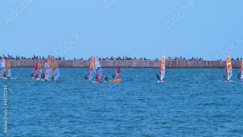 View of young windsurfers participate in local competition, colorful sails, recreational water sports, sunny summer day, calm Baltic sea Karosta beach bay in Latvia, concrete pier, medium distant shot photo