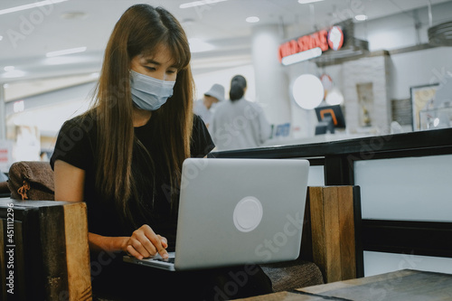 Close up portrait of young blonde female in a medical mask typing keyboard and using laptop computer in coffee shop.