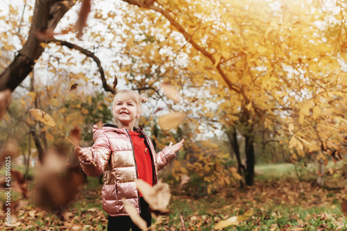 Little blonde girl plays with yellow autumn leaves in the garden, smile, has fun