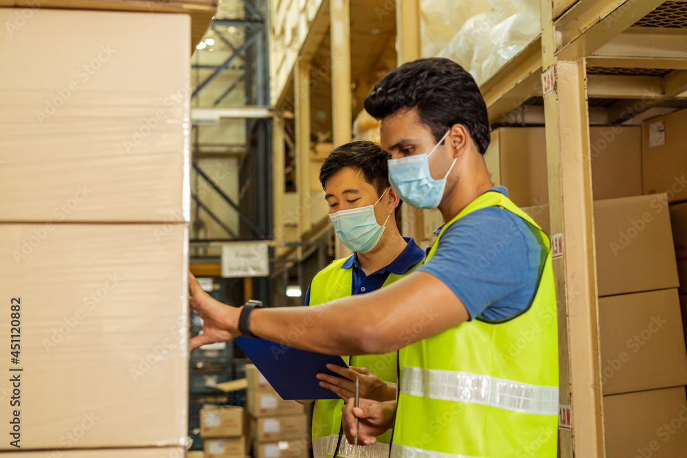 Group of young factory warehouse workers wearing a protective face mask while working in logistic industry indoor. Asian and Indian ethnic men checking item order during Coronavirus Covid 19 pandemic Group of young factory warehouse workers wearing a protective face mask while working in logistic industry indoor. Asian and Indian ethnic men checking item order during Coronavirus Covid 19 pandemic