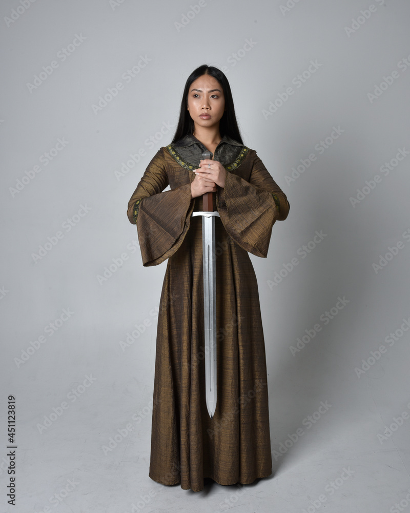 Full length  portrait of beautiful young asian woman with long hair wearing medieval fantasy gown costume. Graceful standing posing holding a sword,  isolated on studio background.
