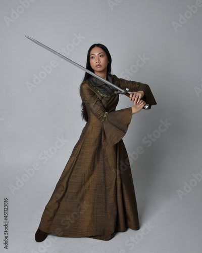 Full length portrait of beautiful young asian woman with long hair wearing medieval fantasy gown costume. Graceful standing posing holding a sword, isolated on studio background.