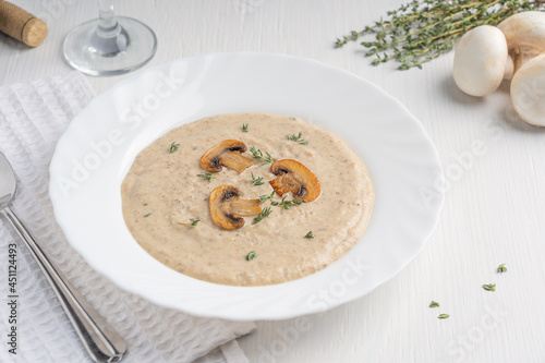 Creamy mushroom soup puree made of blended champignon and dairy cream decorated with fried slices and thyme served for lunch on plate with spoon and glass of wine on white wooden table with textile