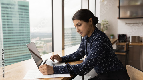 Serious Indian student woman sit at table use laptop do research, studying makes assignment prepare for college admission. Businesswoman making corrections in document, work sit in homeoffice kitchen photo