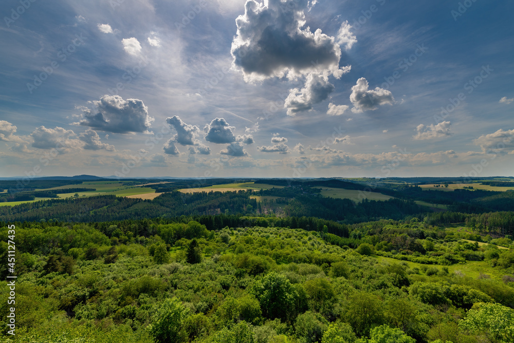 Breathtaking view of a lush green landscape with rolling hills and dense forests under a dramatic sky with scattered clouds. The sun’s rays peek through, casting light over the peaceful countryside.