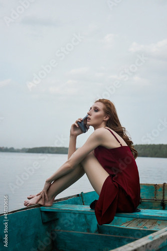 Beautiful fashion redhead woman sit in boat. Beauty romantic portrait girl in red vinous dress in wooden boat on lake © angel_nt