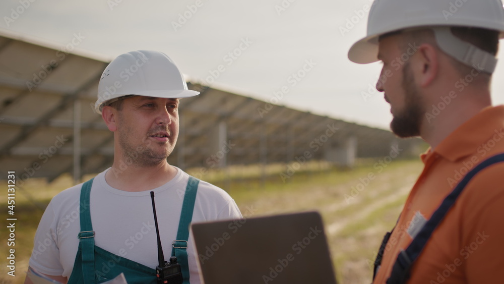 Crop view of diverse male engineers standing at solar farm and discussing efficient plan of construction. Two men in protective helmet and uniform talking and looking at tablet screen. Solar park. Crop view of diverse male engineers standing at solar farm and discussing efficient plan of construction. Two men in protective helmet and uniform talking and looking at tablet screen. Solar park.