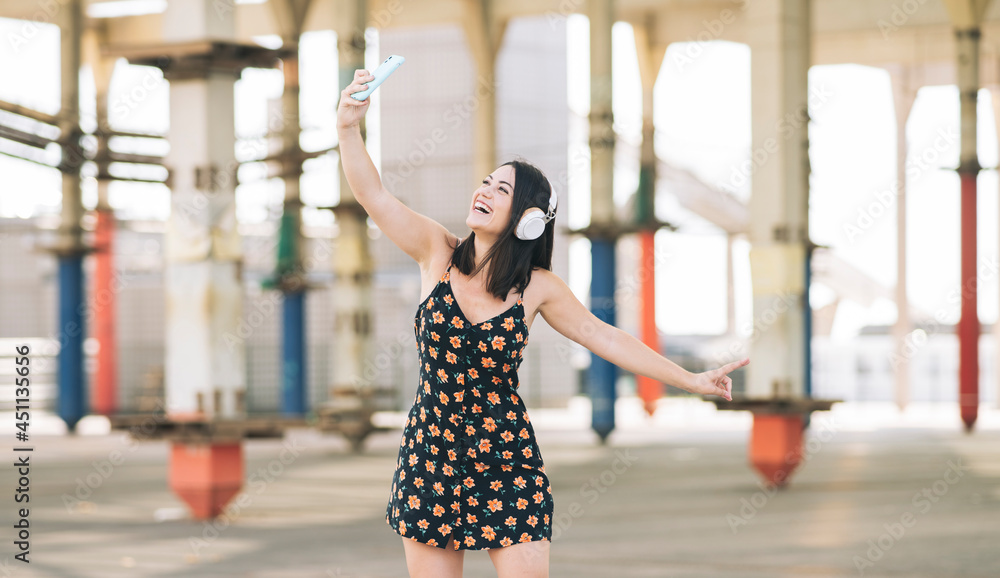 pretty Caucasian woman in a flower dress, dancing listening to music with headphones and with smartphone around the city