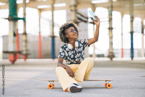 pretty blonde afro woman in glasses with skateboard, sitting takes a photo with smartphone