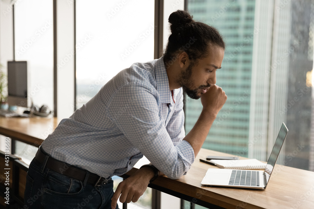 Focused concentrated young African businessman analyst lean over laptop standing alone in modern office working using computer, learning online data, watch webinar thinking of problem solution concept Focused concentrated young African businessman analyst lean over laptop standing alone in modern office working using computer, learning online data, watch webinar thinking of problem solution concept