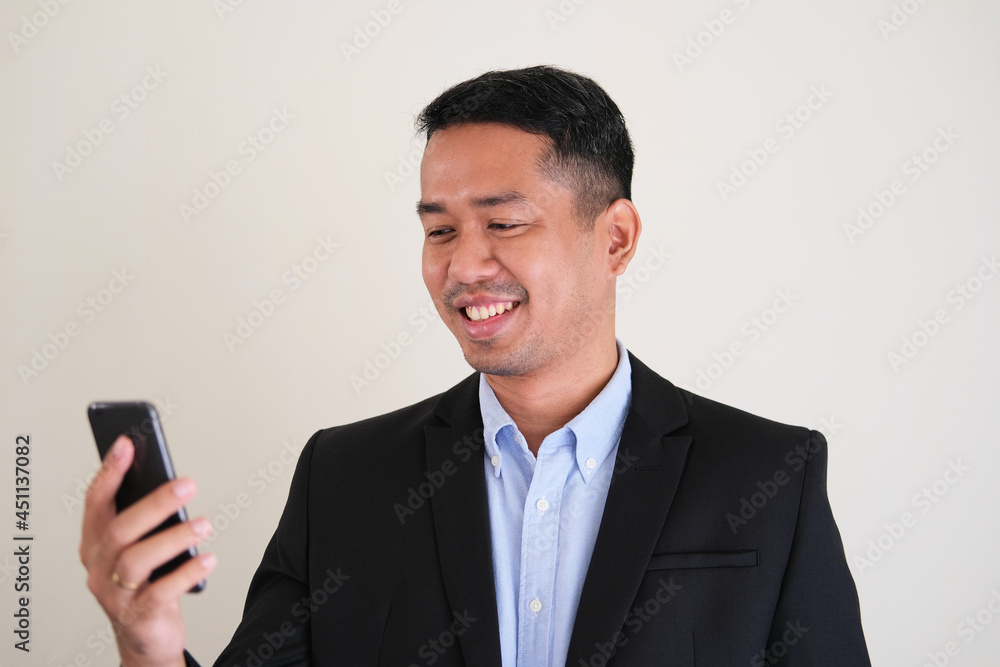 Asian man wearing black suit smiling happy while looking to his mobile phone Asian man wearing black suit smiling happy while looking to his mobile phone