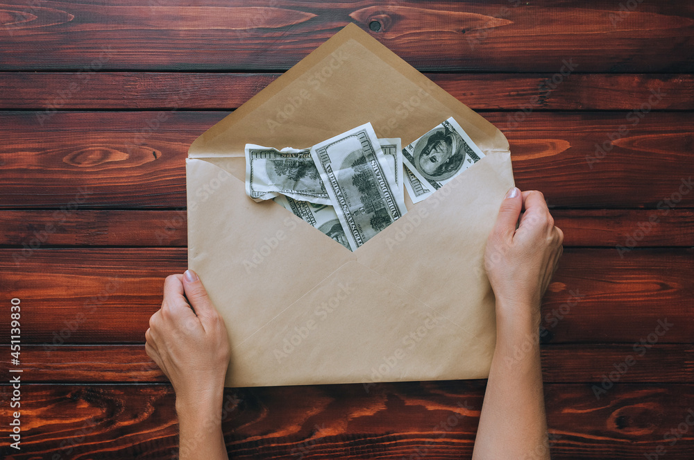 Female hands hold an open beige craft envelope with dollars on a brown wooden background. The concept of donation, alimony, winnings and cashback. View from above. Female hands hold an open beige craft envelope with dollars on a brown wooden background. The concept of donation, alimony, winnings and cashback. View from above.