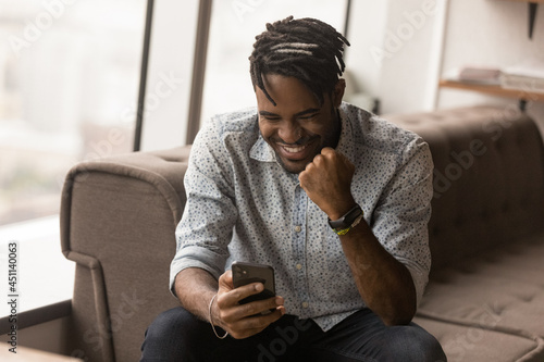 Slika na platnu African hipster guy in dreadlocks hairstyle sit on sofa in modern office read sms on cell phone feels happy celebrate on-line auction betting victory