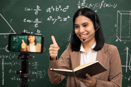 Young asian teacher woman standing looking at camera and video conference with student. Female teacher training the mathematics in classroom from live stream with smartphone online course.