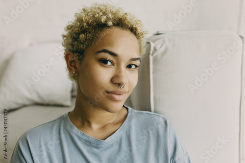 Horizontal close-up picture of curly-headed Afro female with youthful appearance having dyed blonde stylish haircut wearing nose ring and cute long earrings, looking at camera with beautiful brown eye photo