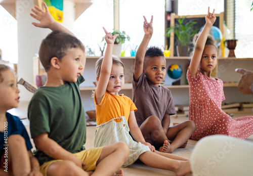 Fotografia Group of small nursery school children sitting on floor indoors in classroom, raising hands