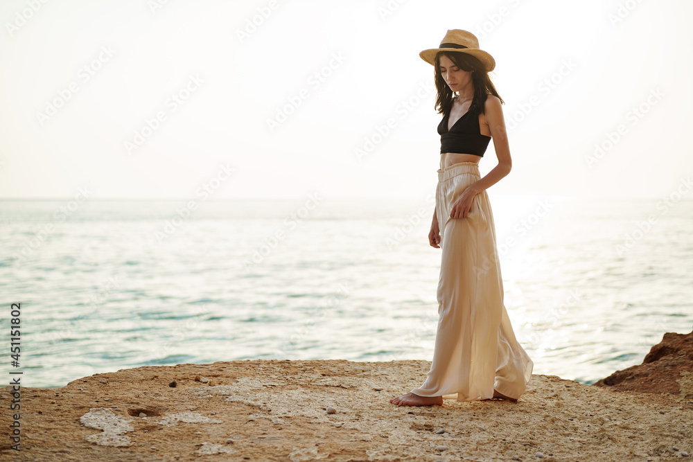 Young stylish woman standing on cliff at the sea at sunset Young stylish woman standing on cliff at the sea at sunset