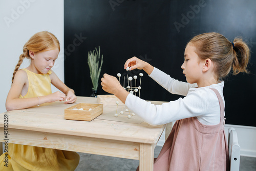 Immersed girls are busy building 3d shapes from plasticine and toothpicks. They all sitting behind the same small square table. Girls wear nice long concervative dresses. photo