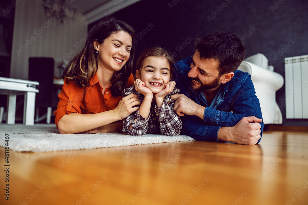 Family lying down on stomach on the floor. Beloved daughter is laughing while parents showing her lots of love. Family lying down on stomach on the floor. Beloved daughter is laughing while parents showing her lots of love.