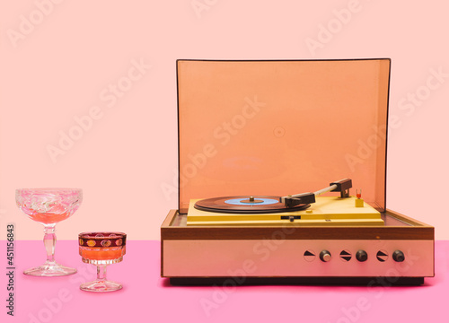 Gramophone record player from sixties. Turntable for single vinyl with pink bottom with light pink background. Two cocktail glasses, party vibe.