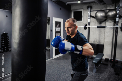 Energy and concept boxing. A dedicated man wears boxing gloves and sports clothes in an indoor modern gym with equipment. Man in sports and boxing is just getting ready to hit a big black punching bag