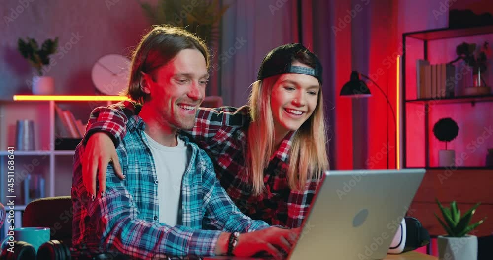 Lovely happy loving smiling blond girl hugging and kissing to the cheek her positive boyfriend when they together making purchases in online market on computer at home with night lighting Lovely happy loving smiling blond girl hugging and kissing to the cheek her positive boyfriend when they together making purchases in online market on computer at home with night lighting