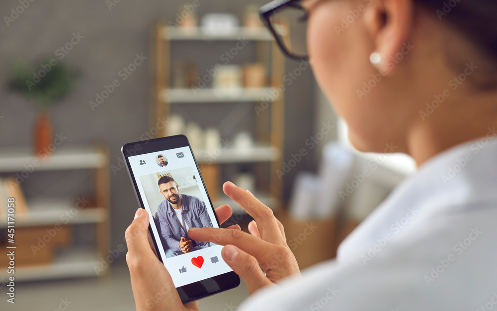 Woman looking for romantic relationship. Single lady holding smartphone and giving like to photo of young man on online dating mobile app or website. Closeup display shot, close up view over shoulder