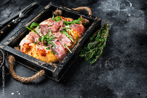 Italian Pizza with prosciutto parma ham, arugula salad and cheese in a rustic wooden tray. Black background. Top view. Copy space