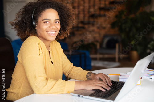 Happy young African-American woman wearing headset using laptop for remote work or studying, sitting at the home office and looks at the camera, sales woman making calls to customers