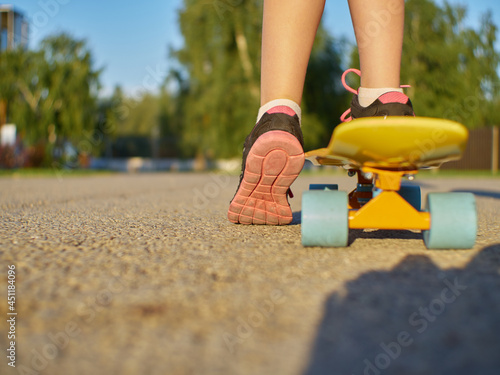 Girl kid standing on the asphalt road and yellow penny board with blue wheels on street background during sunset time.
