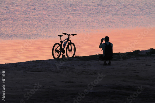 Man beside the bike takes a picture of the sunset near the water