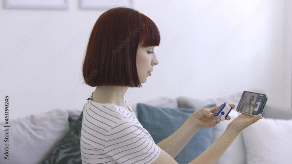 Doctor online connection. Young woman sits on sofa at home with pulse oximeter, talking with male professional doctor in white coat and hijab over internet video connection Doctor online connection. Young woman sits on sofa at home with pulse oximeter, talking with male professional doctor in white coat and hijab over internet video connection
