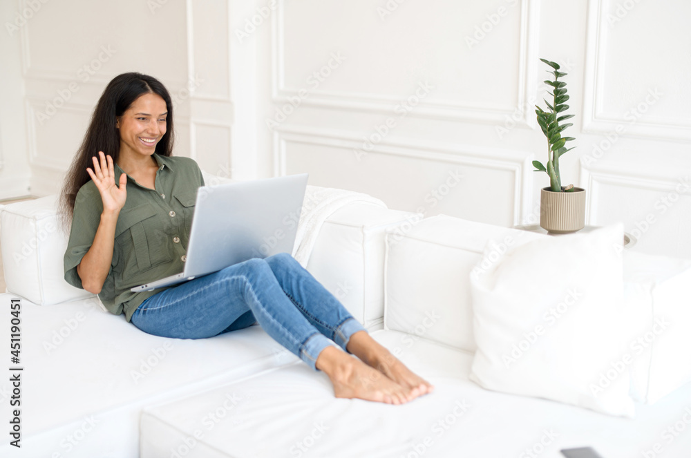 Happy smiling indian woman with a laptop has video call, virtual meeting. A young mixed-race female lying down on the white comfortable couch and waving into webcam cheerfully Happy smiling indian woman with a laptop has video call, virtual meeting. A young mixed-race female lying down on the white comfortable couch and waving into webcam cheerfully