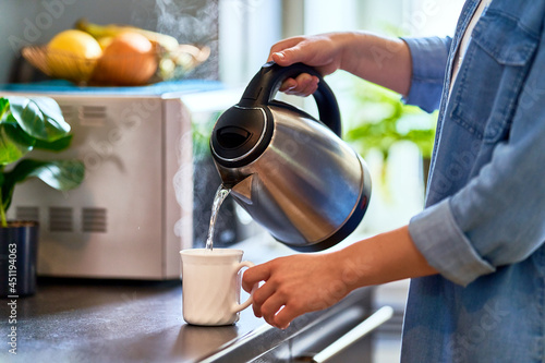 Female hands pouring boiling water from a modern metal stainless kettle in a glass cup for brewing tea in the kitchen at home photo