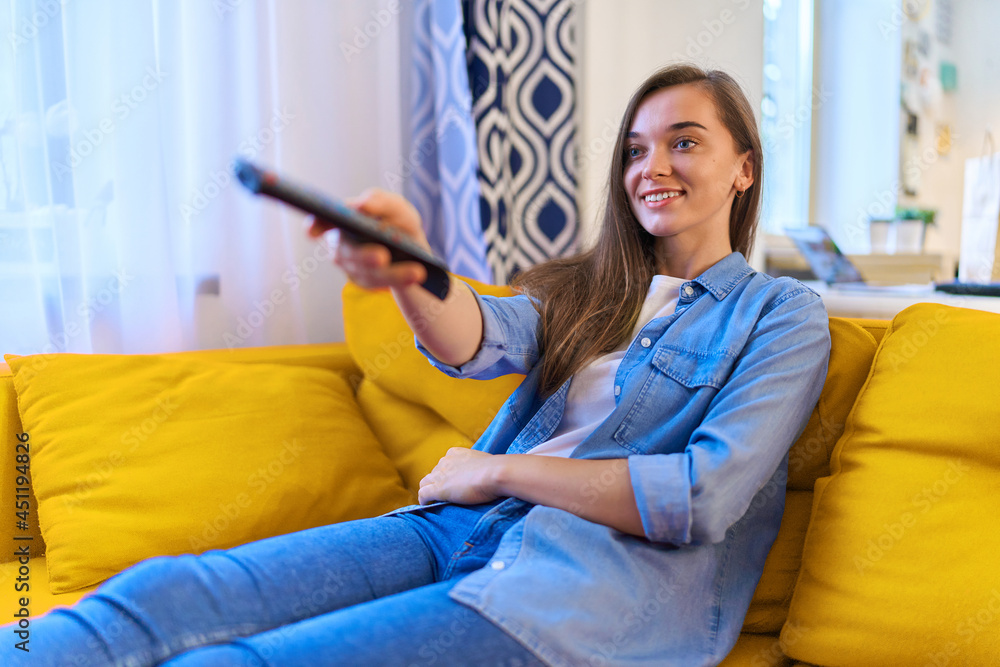 Portrait of happy young satisfied smiling attractive girl sitting on sofa, switches channels on the remote control and watching tv at home alone Portrait of happy young satisfied smiling attractive girl sitting on sofa, switches channels on the remote control and watching tv at home alone