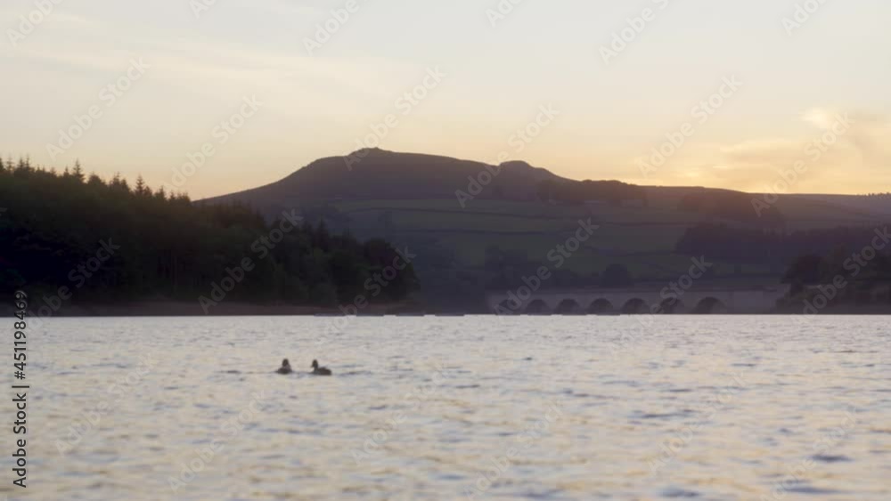Handheld static shot of silhouette of two ducks swimming in Ladybower Reservoir, Upper Derwent Valley, Derbyshire, England at sunset. Ashopton Viaduct, forest and mountains visible in background. Handheld static shot of silhouette of two ducks swimming in Ladybower Reservoir, Upper Derwent Valley, Derbyshire, England at sunset. Ashopton Viaduct, forest and mountains visible in background.