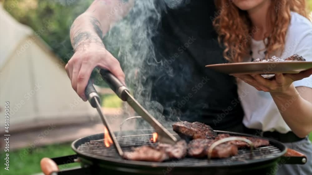 Young couple frying meat on the grill. Greenery around. Glamping. Slow motion Young couple frying meat on the grill. Greenery around. Glamping. Slow motion