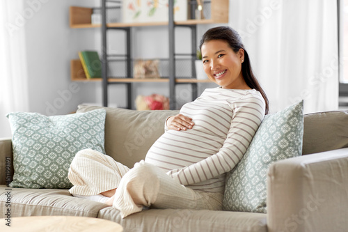 pregnancy, rest, people and expectation concept - happy smiling pregnant asian woman sitting on sofa at home