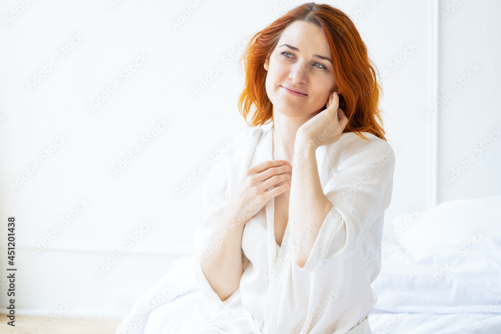 Beauty portrait smiling ginger woman middle age in silk robe doing a facial massage in light room Beauty portrait smiling ginger woman middle age in silk robe doing a facial massage in light room