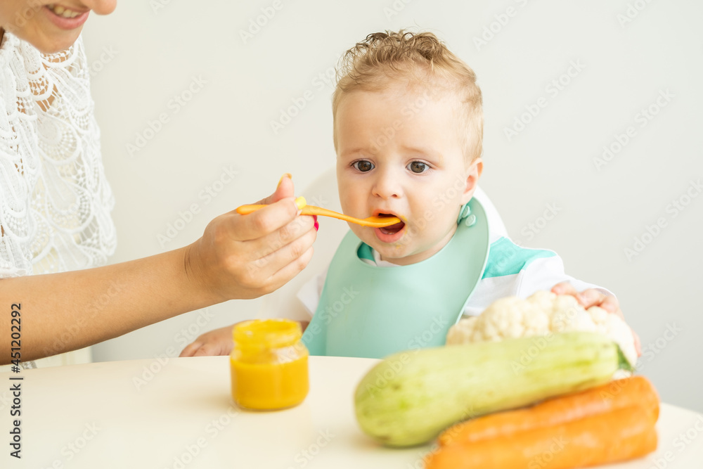 Baby boy sitting in a Childs chair eating vegetable puree on white kitchen. Mom feeds baby. Baby boy sitting in a Childs chair eating vegetable puree on white kitchen. Mom feeds baby.