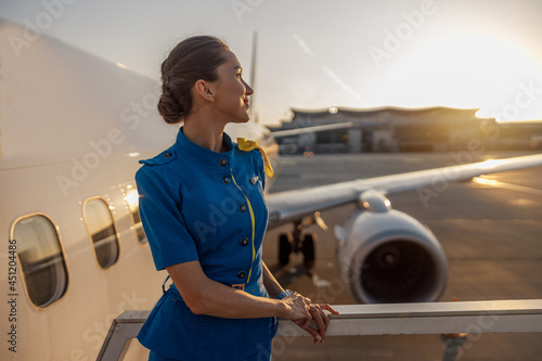 Thoughtful air stewardess in blue uniform looking away, standing outdoors at the sunset. Commercial airplane near terminal in an airport in the background. Aircrew, occupation concept photo
