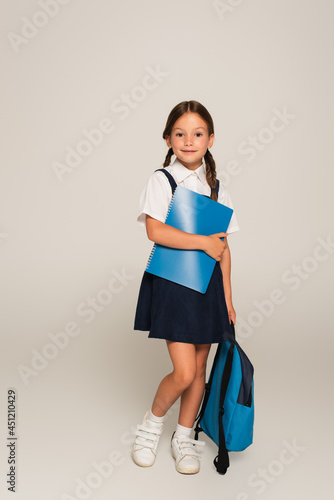 Fotografía full length view of smiling schoolgirl standing with blue backpack and copy book