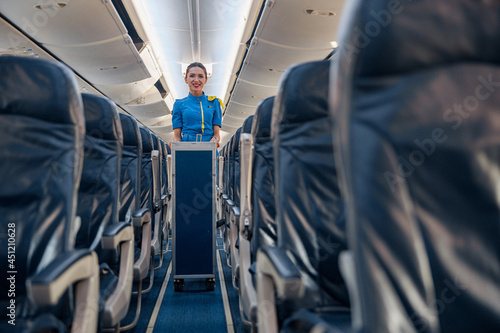 Female cabin attendant leading trolley cart through empty plane aisle. Travel, service, transportation, airplane concept