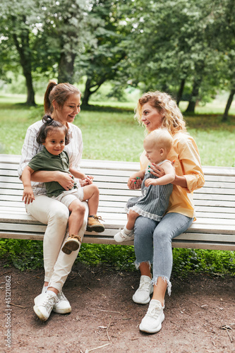 Full length portrait of two young mothers sitting on bench in park and playing with cute babies