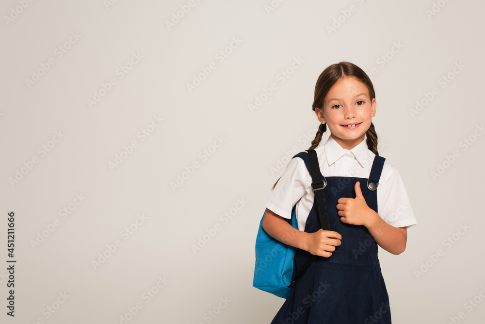 happy schoolgirl with blue backpack showing thumb up isolated on grey happy schoolgirl with blue backpack showing thumb up isolated on grey