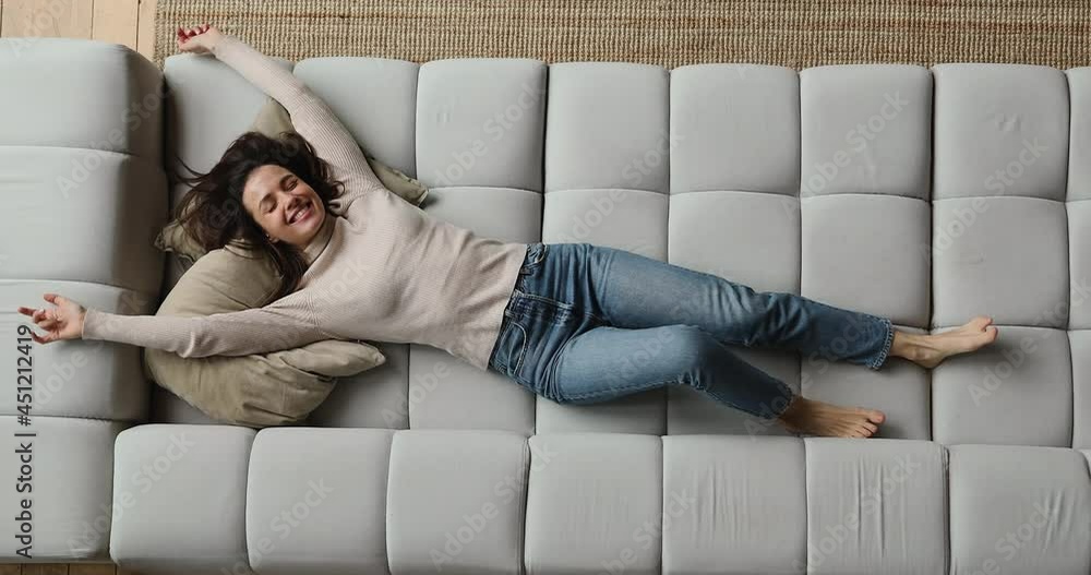 Top above view happy young beautiful smiling woman falling on huge comfortable sofa, raising arms stretching back muscles, enjoying carefree peaceful weekend relaxation time alone in living room.