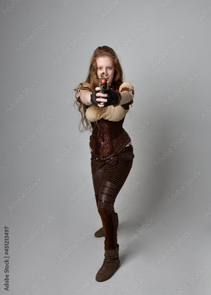 Full length portrait of beautiful young woman with long red hair, wearing steampunk inspired costume.  Posing while holding a gun,  isolated on studio background.